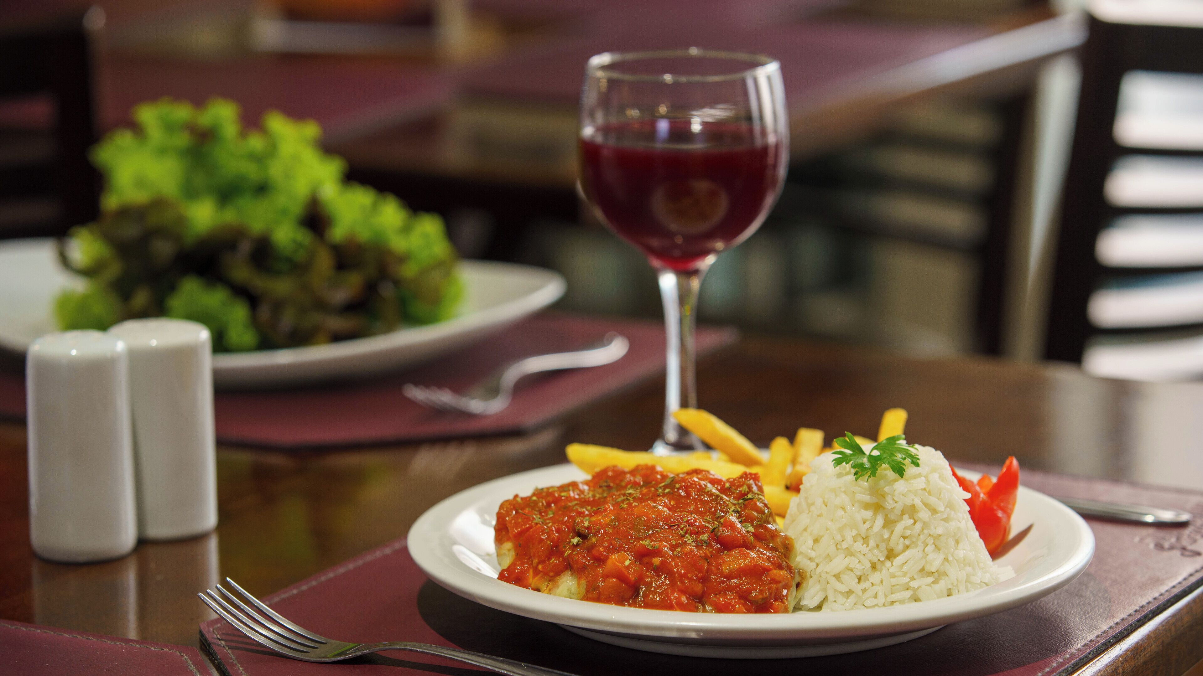 Un plato de comida con arroz, un guiso de tomate y papas fritas, acompañado de una copa de vino tinto, sobre una mesa con salero y pimentero.