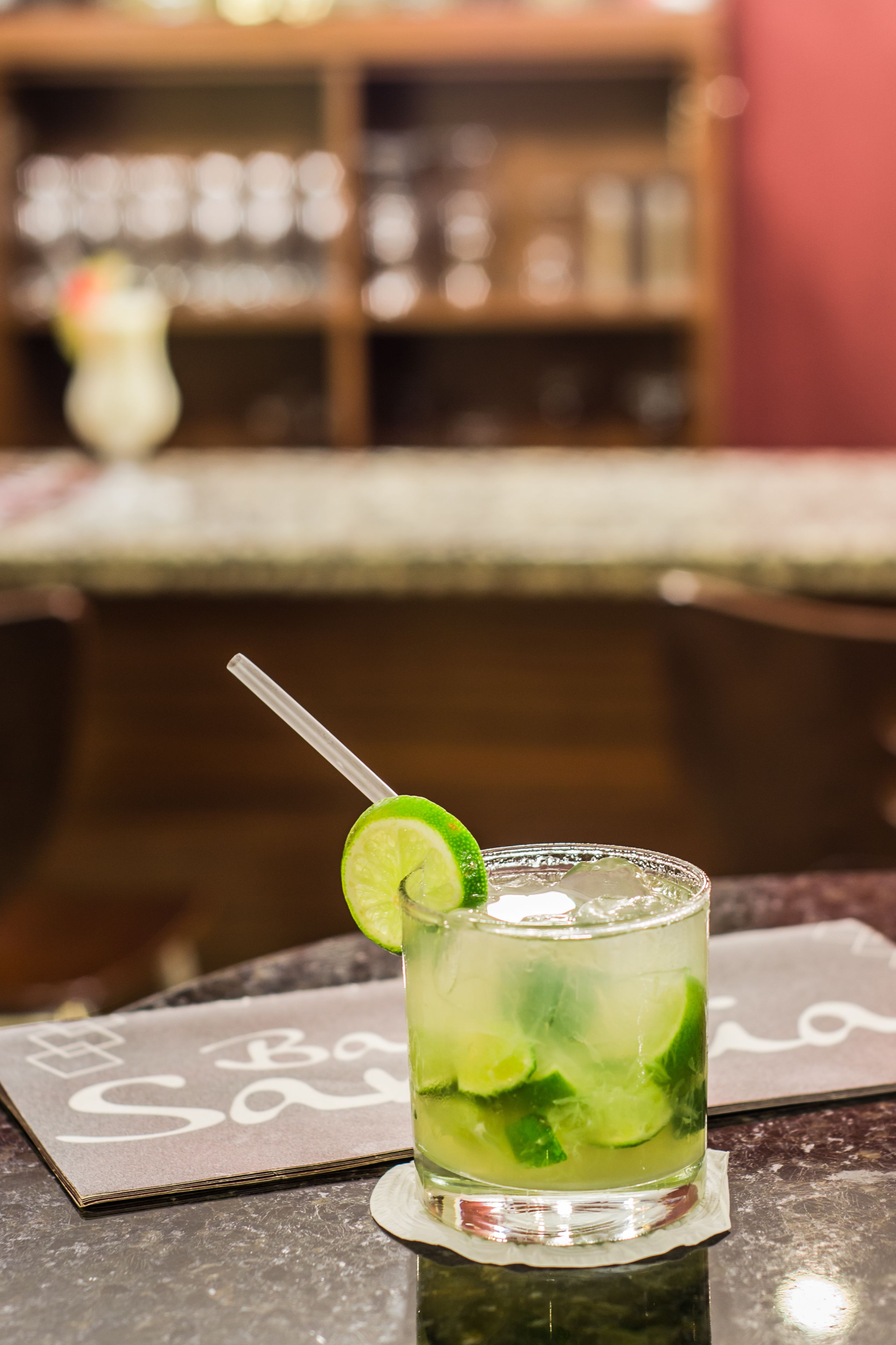 Un vaso con un cóctel verde y una cuña de lima sobre un posavasos, colocado sobre una mesa con un menú al fondo.