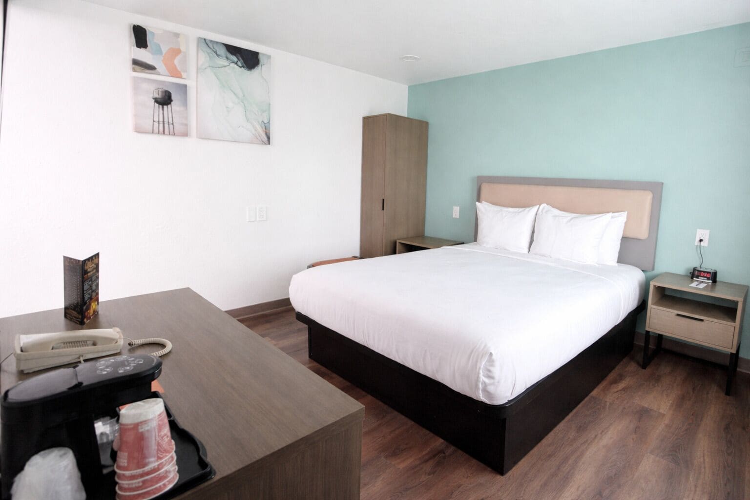 Habitación de hotel con cama, mesitas de noche, escritorio, teléfono y cafetera, además de un cuadro abstracto en la pared.