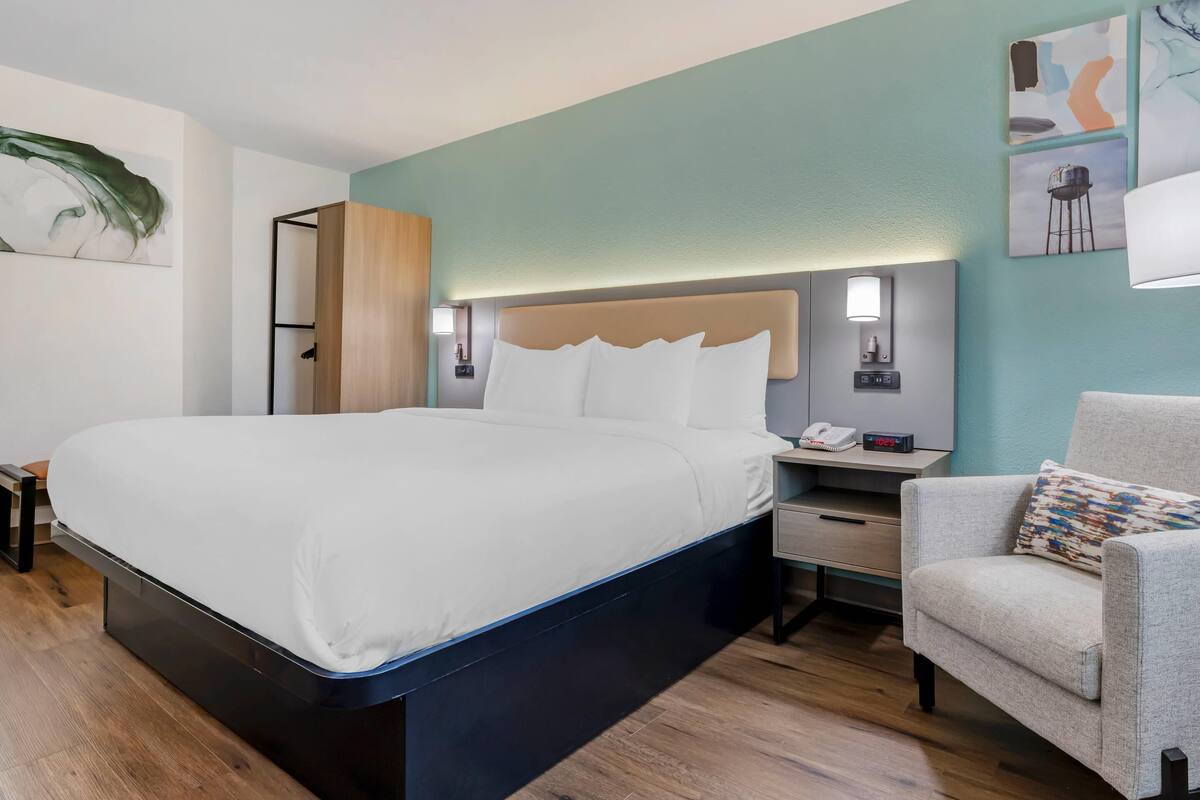 A modern hotel room with a large bed, bedside tables, a chair, and artwork on the walls.