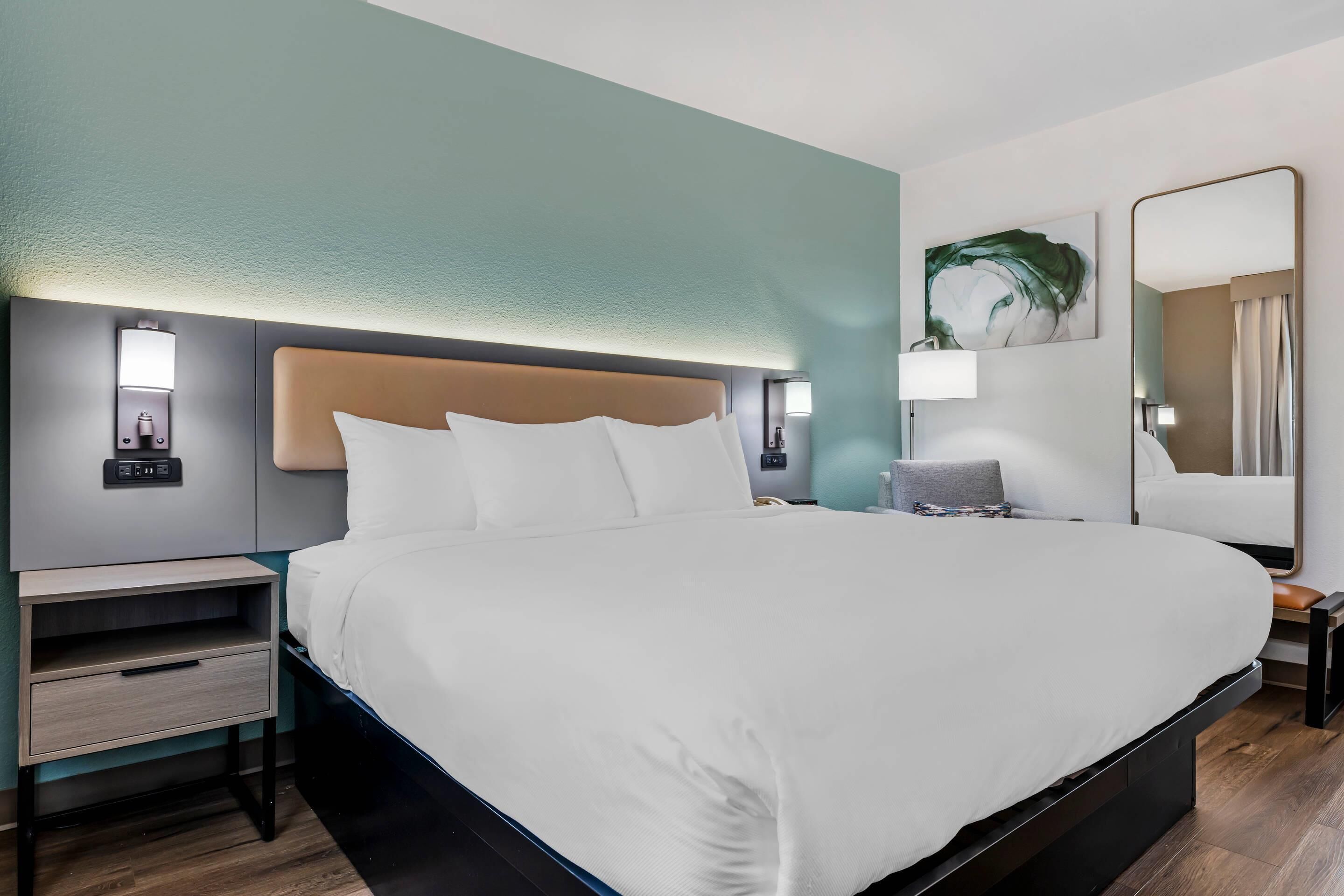 A hotel room with a large bed, bedside tables, a nightstand, a wall-mounted lamp, a mirror, and a painting.