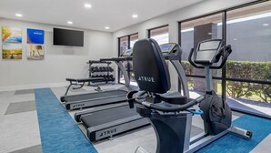 Un gimnasio con cintas de correr, elípticas y mancuernas, con un televisor en la pared y ventanas amplias.