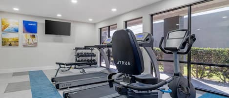 A gym with treadmills, an elliptical, and dumbbells, with a TV on the wall and large windows.