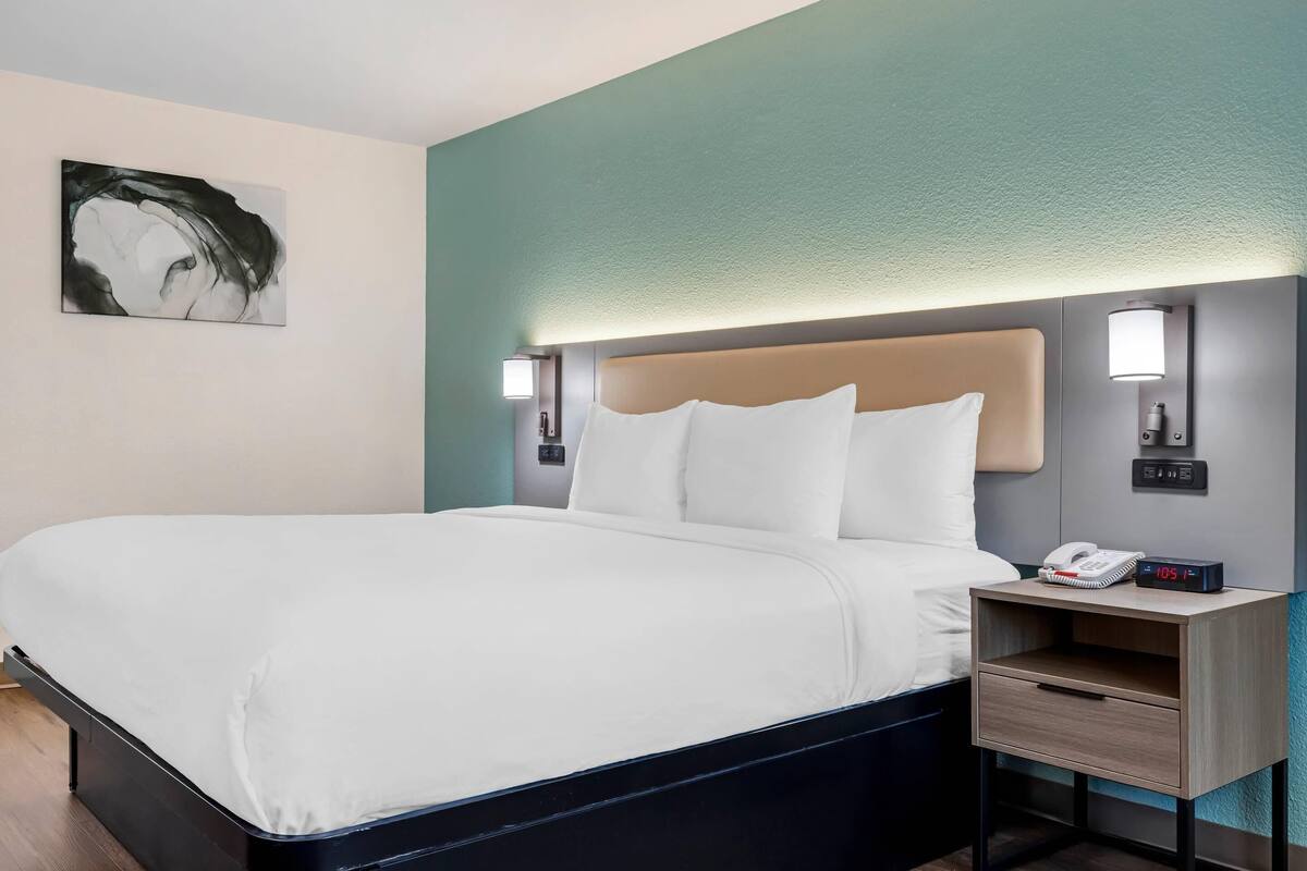 A hotel room with a bed, bedside table, lamp, and artwork on the wall.