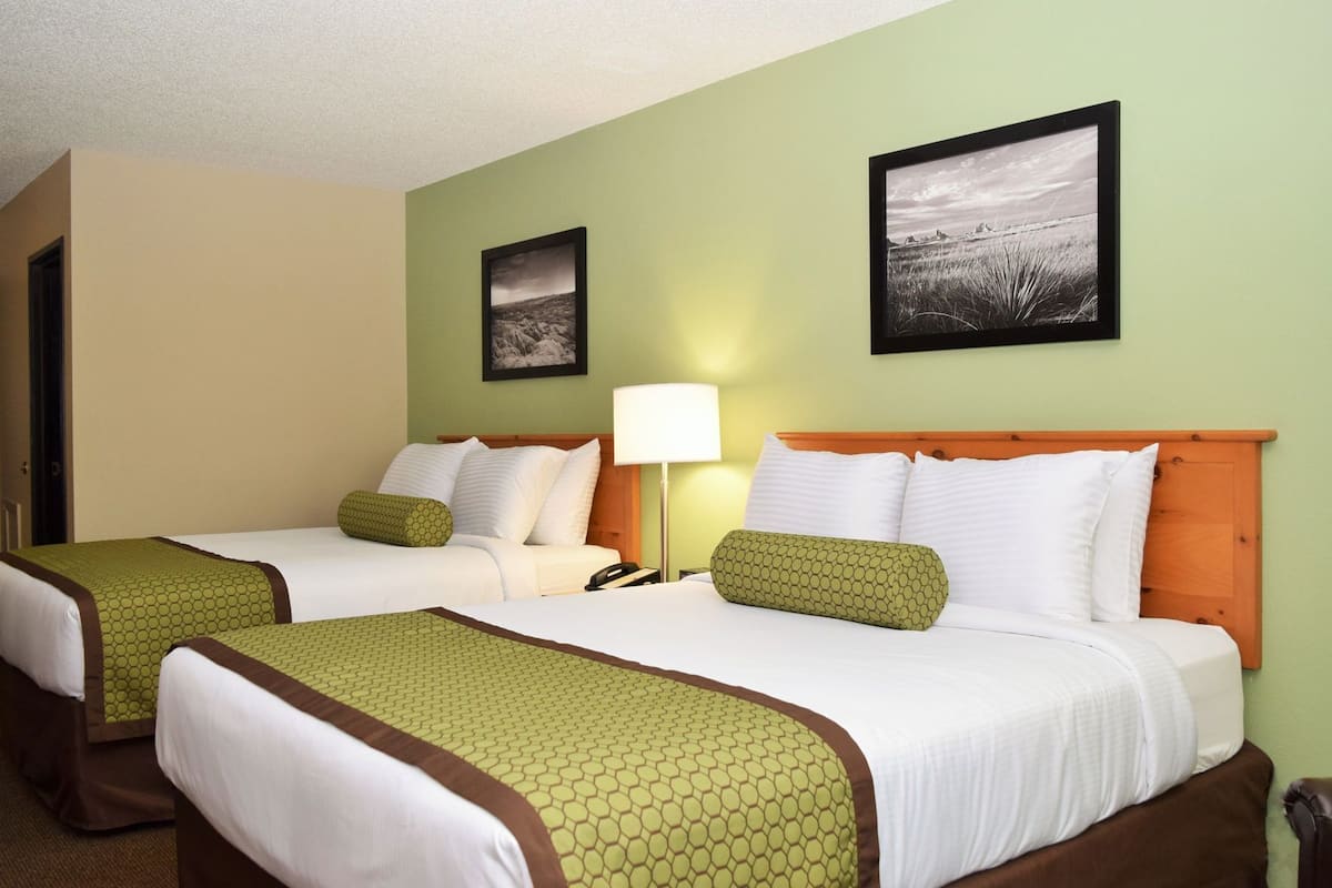 A hotel room with two beds, green and brown bedding, a nightstand with a lamp, and two framed pictures on the wall.