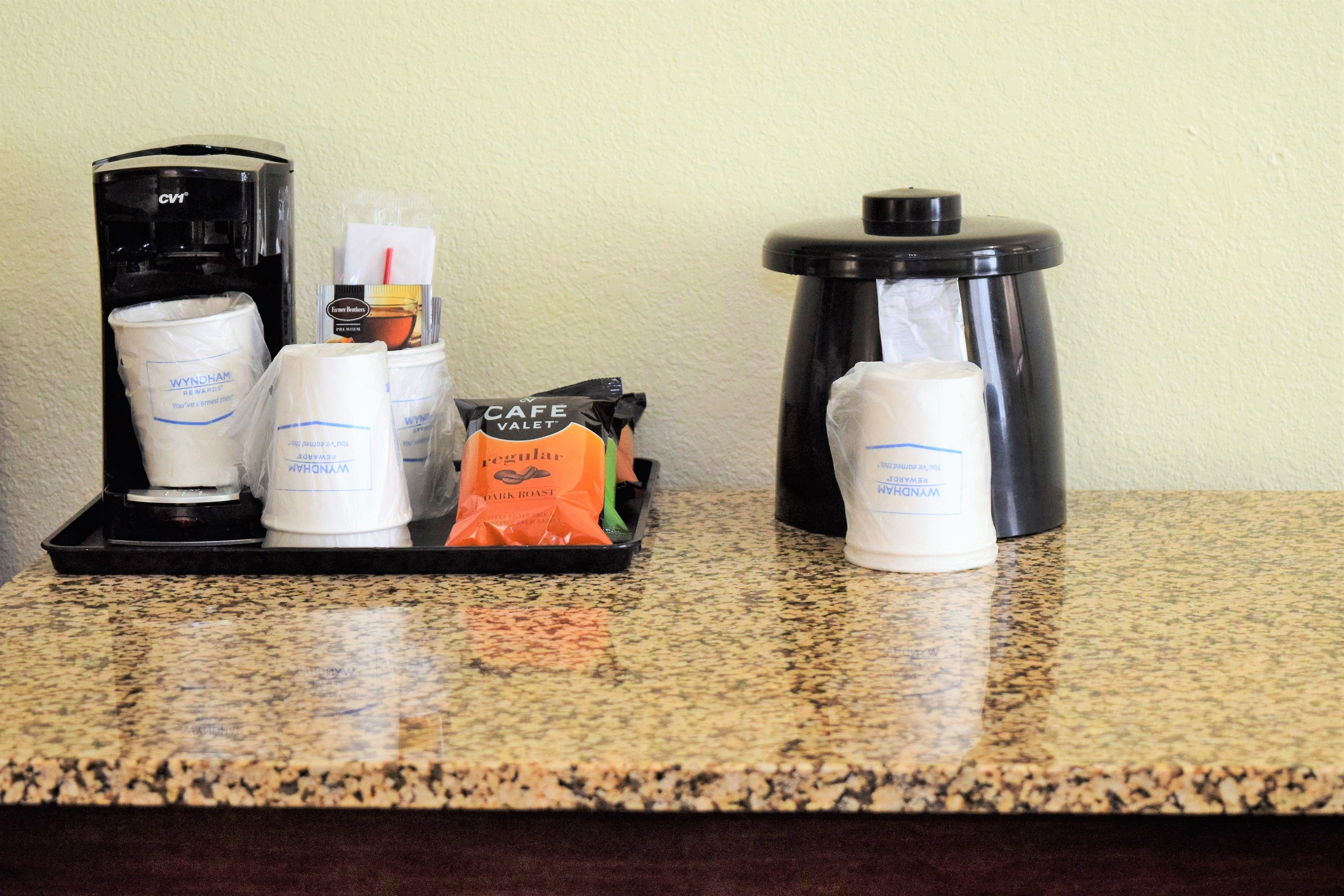 A coffee station with a coffee maker, cups, a bag of coffee pods, and a trash can.