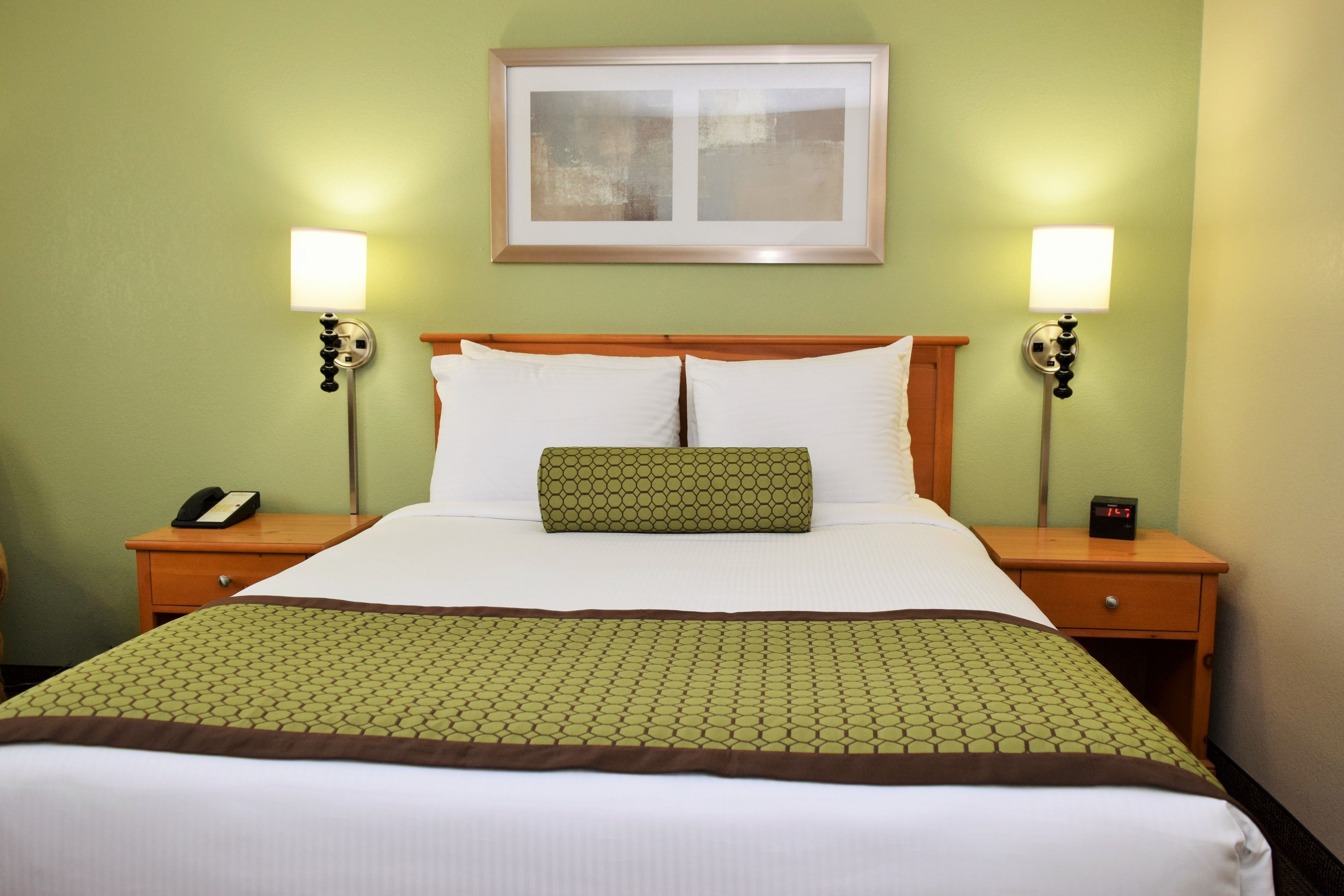 Quarto de hotel com uma cama grande, duas mesas de cabeceira com abajures, um quadro na parede e um edredom verde com padrão.