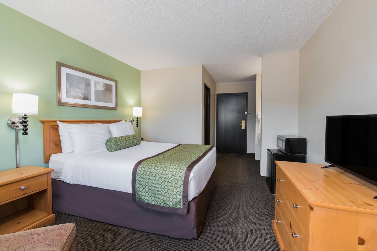 A hotel room with a bed, bedside lamps, a television, a dresser, and a door leading to another room.