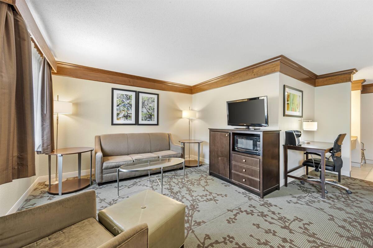 Ein modernes Hotelzimmer mit einer Couch, einem kleinen Tisch, einem Schreibtisch mit Stuhl, einem Fernseher und gerahmten Kunstwerken an den Wänden.