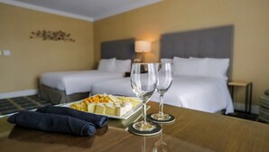 A hotel room with a neatly made bed, a bedside table with a plate of cheese and two wine glasses, and a lamp on the nightstand. - The Grand Hotel (Sunnyvale)