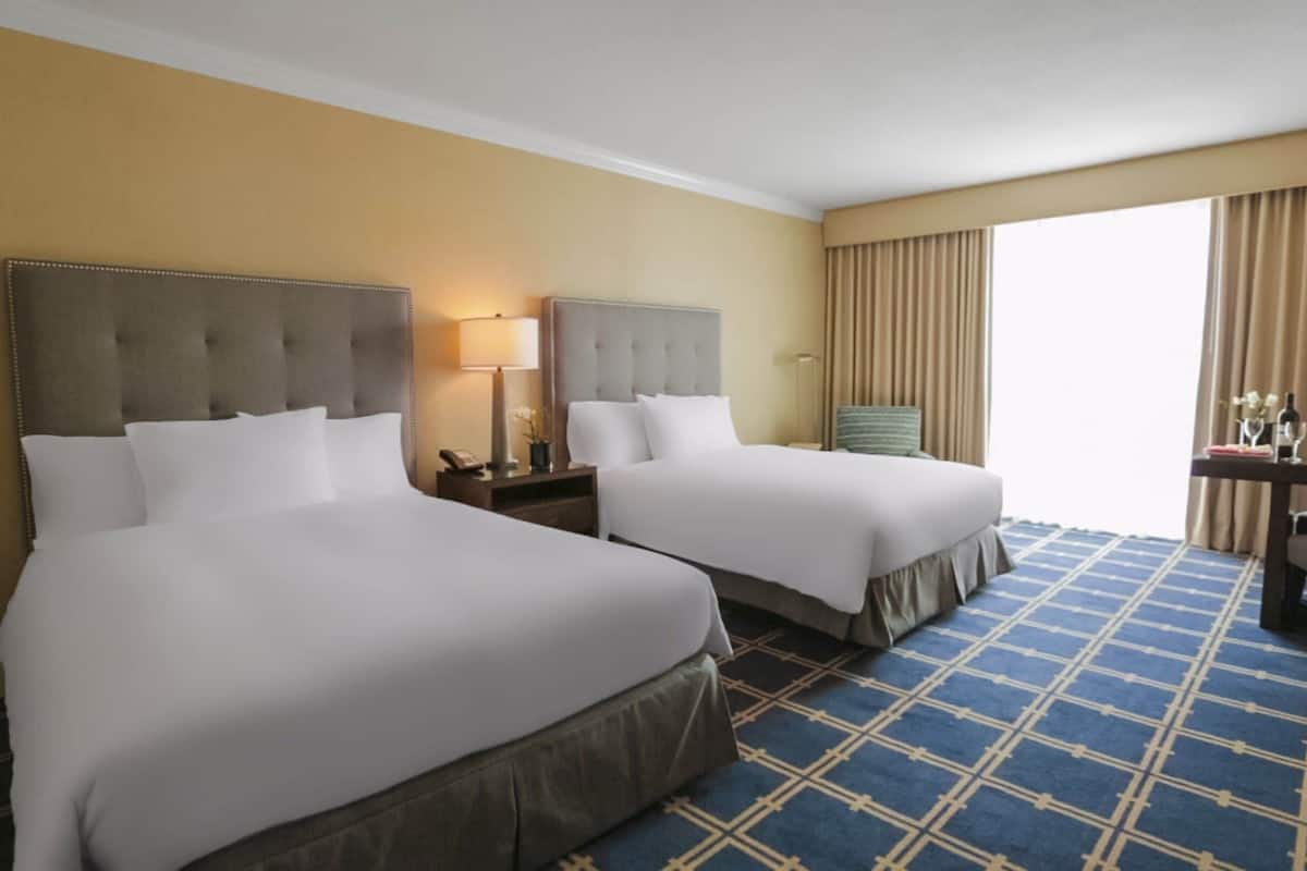 Habitación de hotel con dos camas, un escritorio, una silla y un amplio ventanal con cortinas.