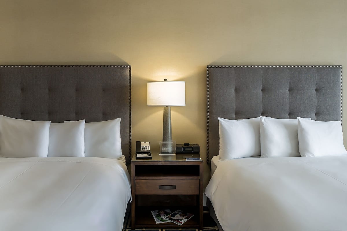 Habitación de hotel con dos camas, cada una con un cabecero tapizado gris, ropa de cama blanca y una mesita de noche con lámpara, teléfono y revistas.