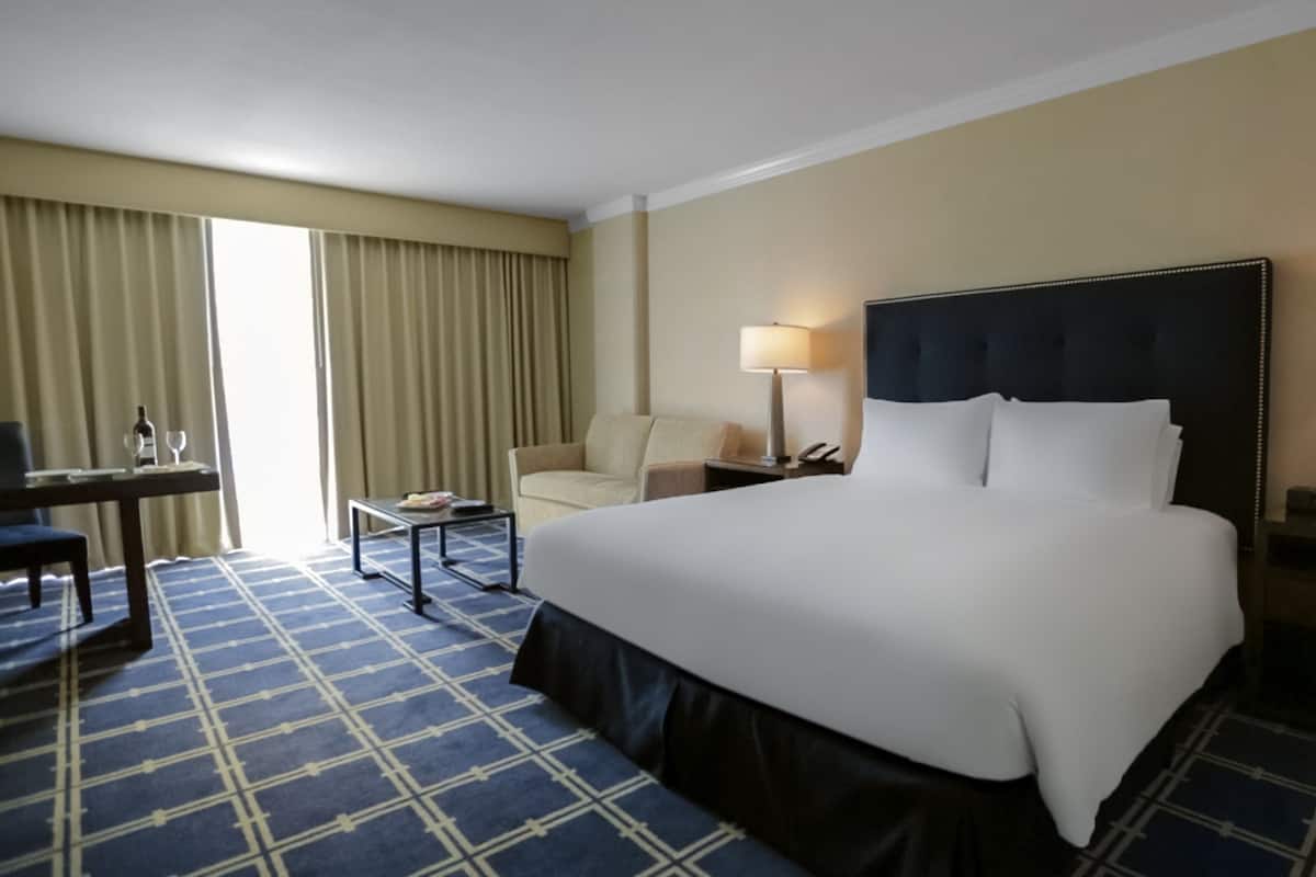Habitación de hotel con una cama grande, un escritorio con silla, una lámpara y una ventana con cortinas.