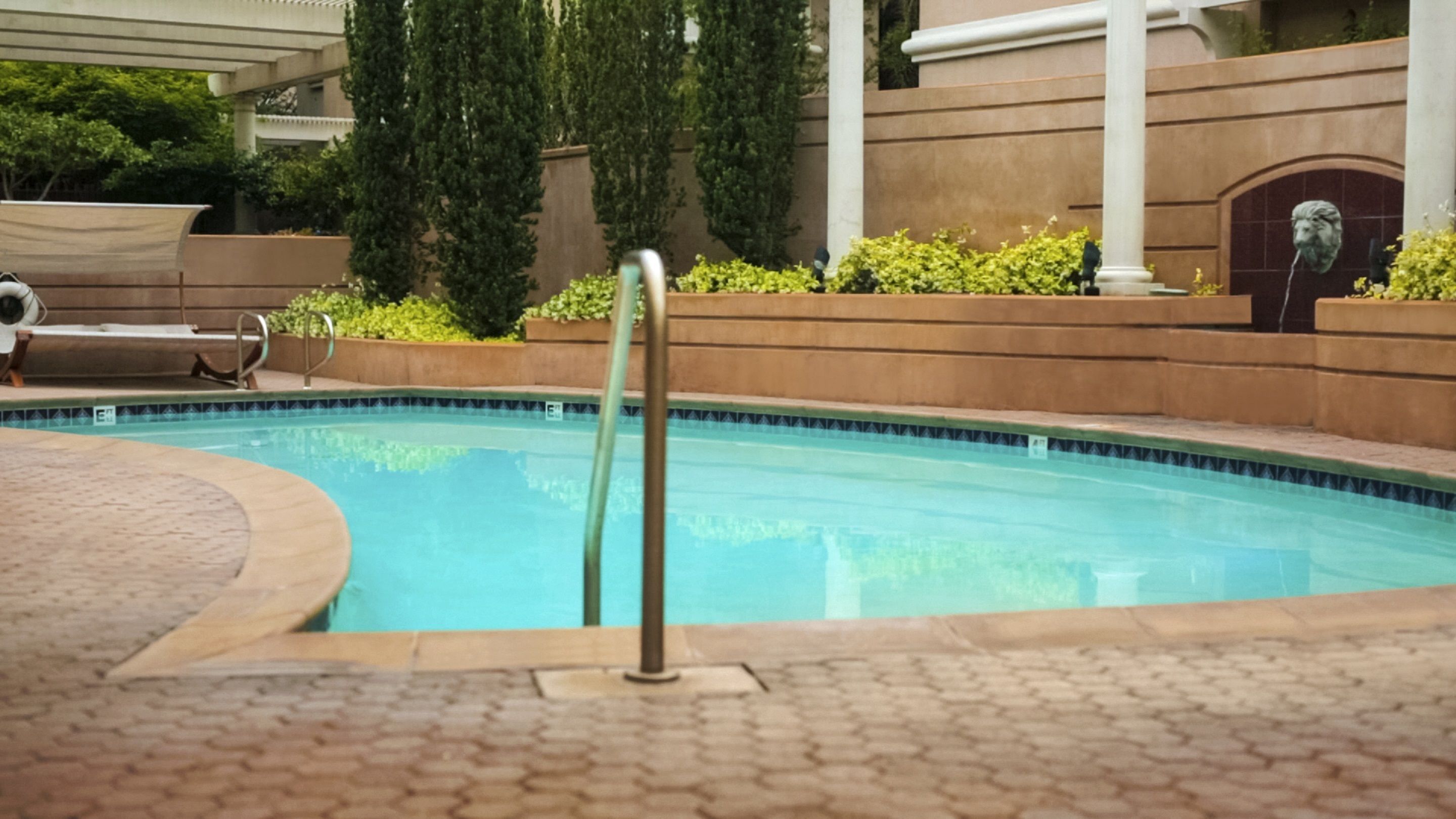 A swimming pool with a metal ladder, surrounded by a tiled deck and greenery.