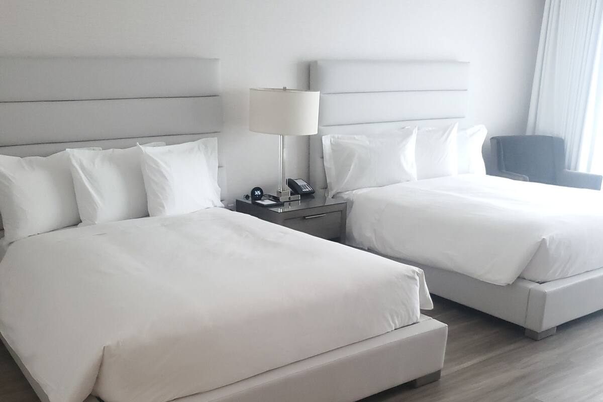 Habitación de hotel con dos camas, una mesita de noche con lámpara y una silla.