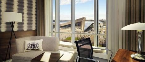 Een hotelkamer met een bank, een kleine tafel, een stoel, een bureau en een lamp. Er is een groot raam met uitzicht op een gebouw en een wateroppervlak.