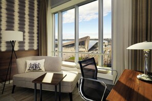 Habitación de hotel con sofá, una mesita, una silla, escritorio y lámpara. Se ve un ventanal con vistas a un edificio y una masa de agua.