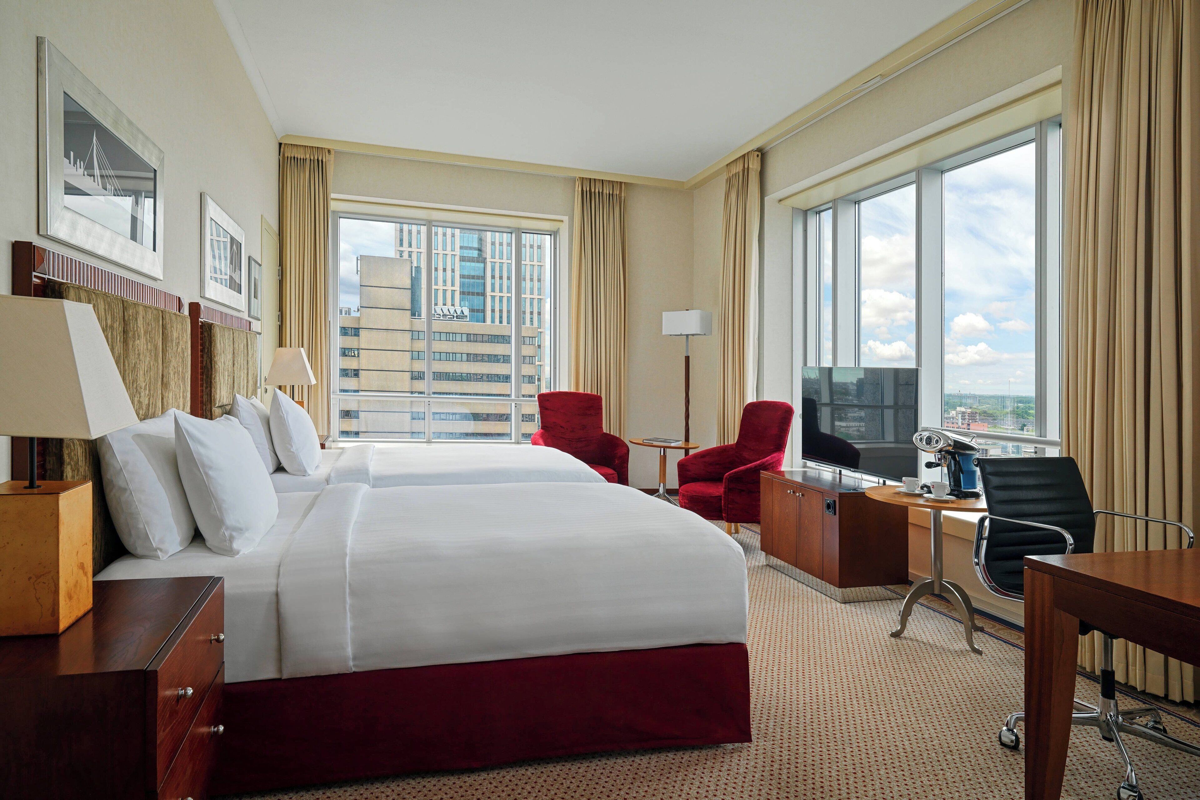 Quarto de hotel com uma cama grande, uma escrivaninha, uma cadeira e vista para a paisagem urbana através de grandes janelas.