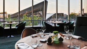 Un restaurante con vistas a un edificio moderno a través de amplios ventanales. Las mesas están dispuestas con copas de vino y cubiertos.