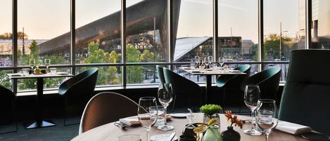 A restaurant with a view of a modern building through large windows. Tables are set with wine glasses and cutlery.