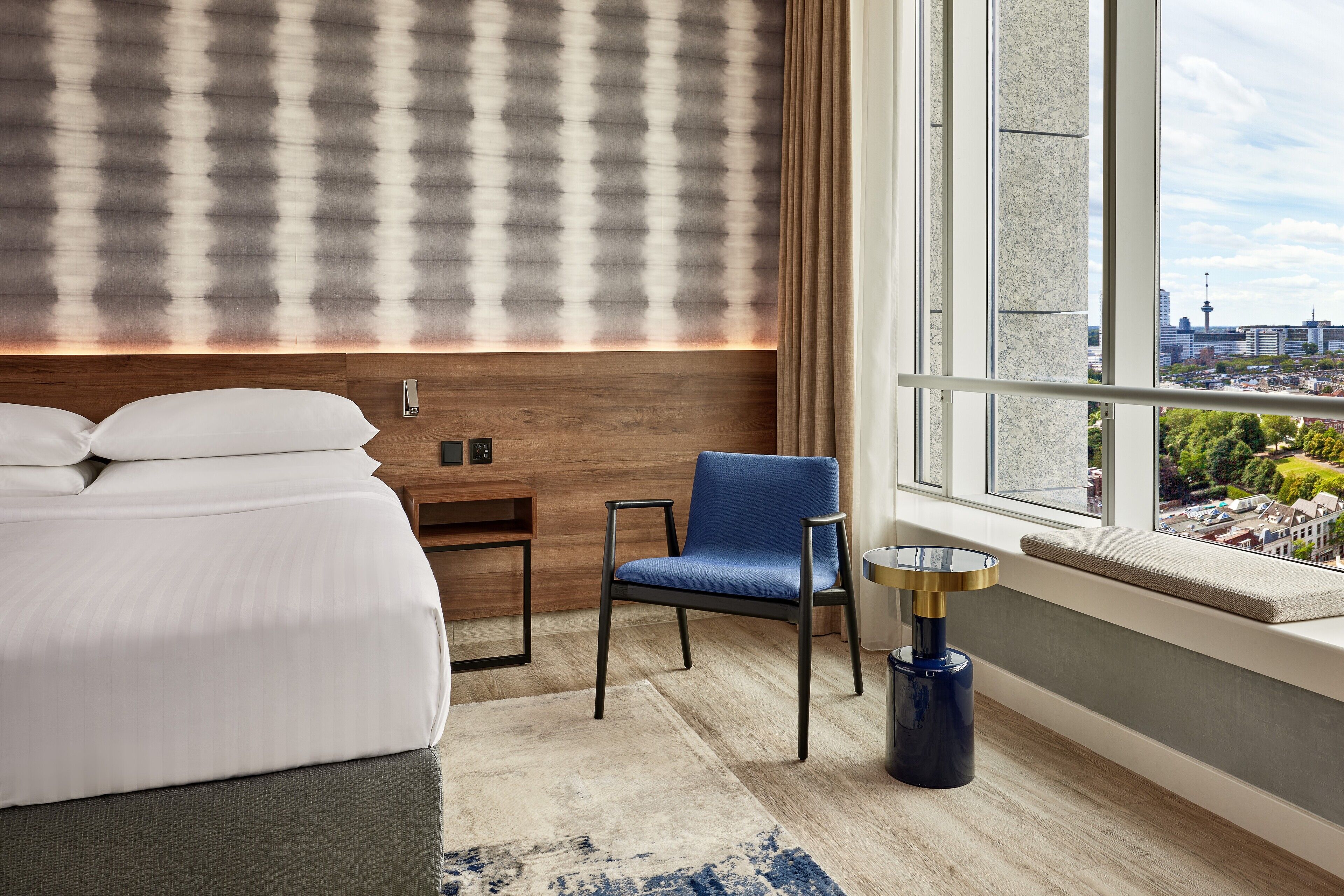 Een moderne hotelkamer met een groot bed, een blauwe stoel, een kleine tafel en uitzicht op de stad door grote ramen.