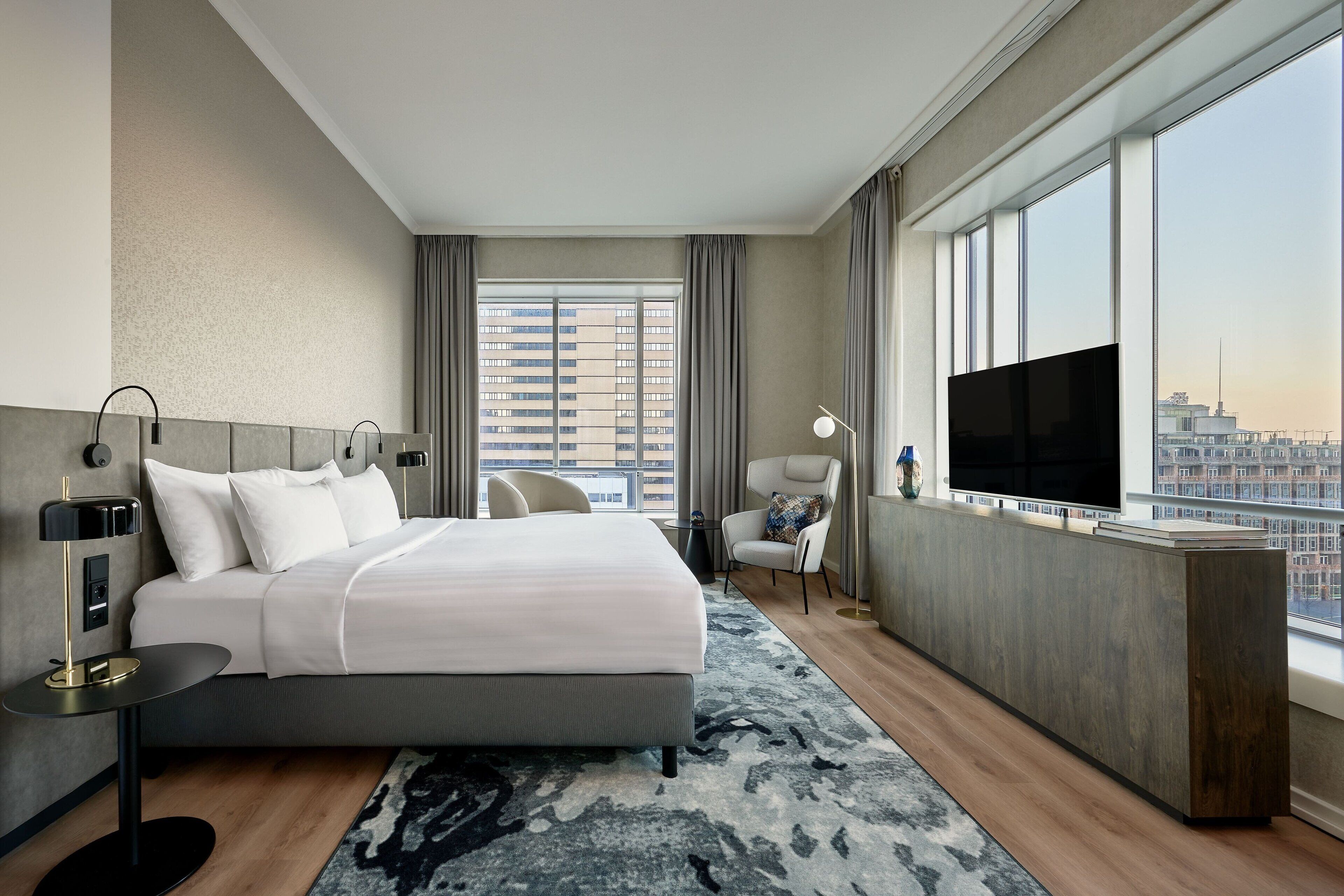 Une chambre d’hôtel moderne dotée d’un grand lit, d’une télévision, d’un bureau et offrant une vue sur la ville grâce à de grandes fenêtres.
