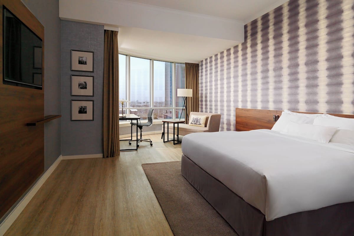 Habitación de hotel con una cama grande, un escritorio y vistas de la ciudad a través de amplias ventanas.
