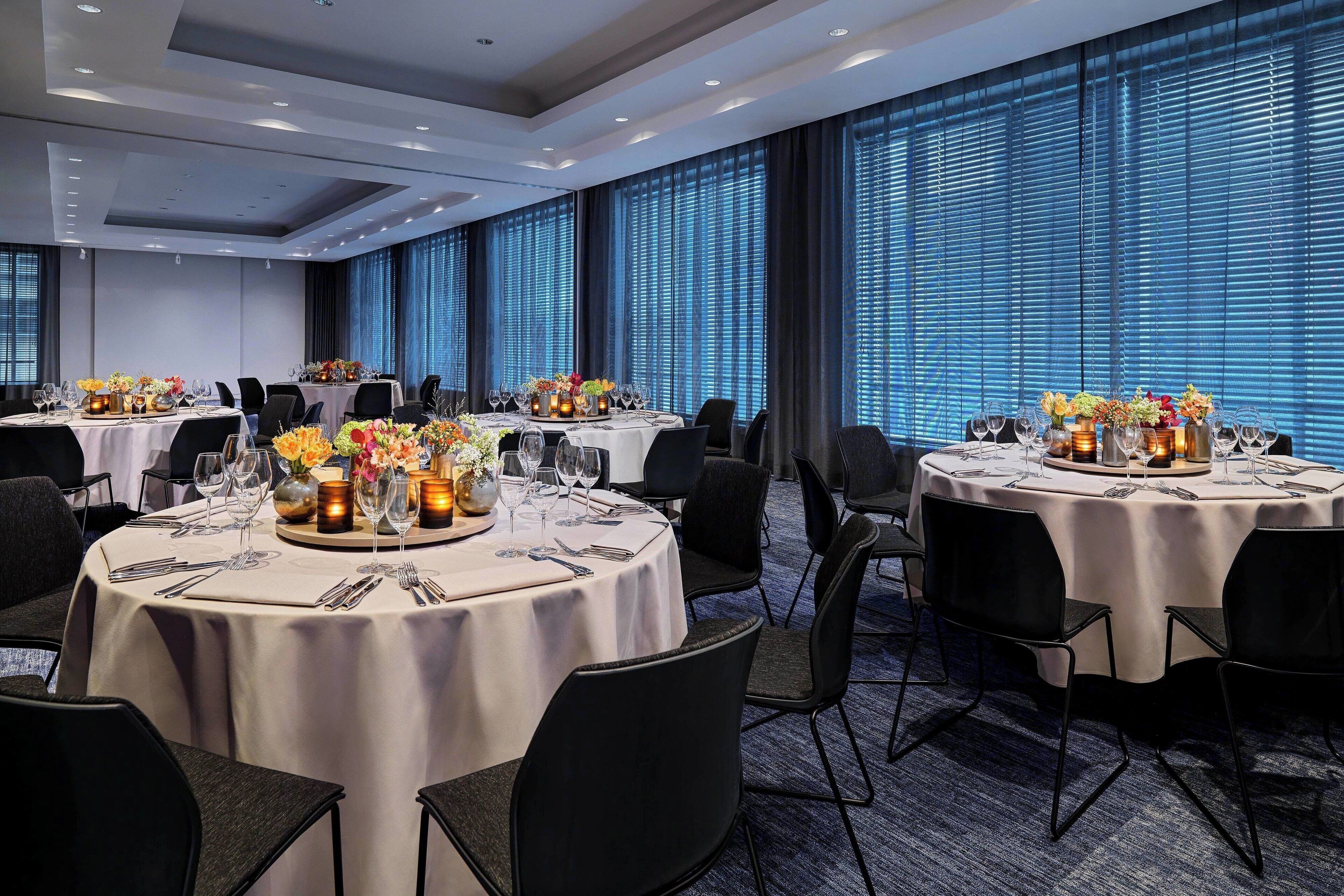 Un comedor de hotel con mesas redondas dispuestas para seis comensales, cada una con mantel blanco, sillas negras y centros de mesa florales.