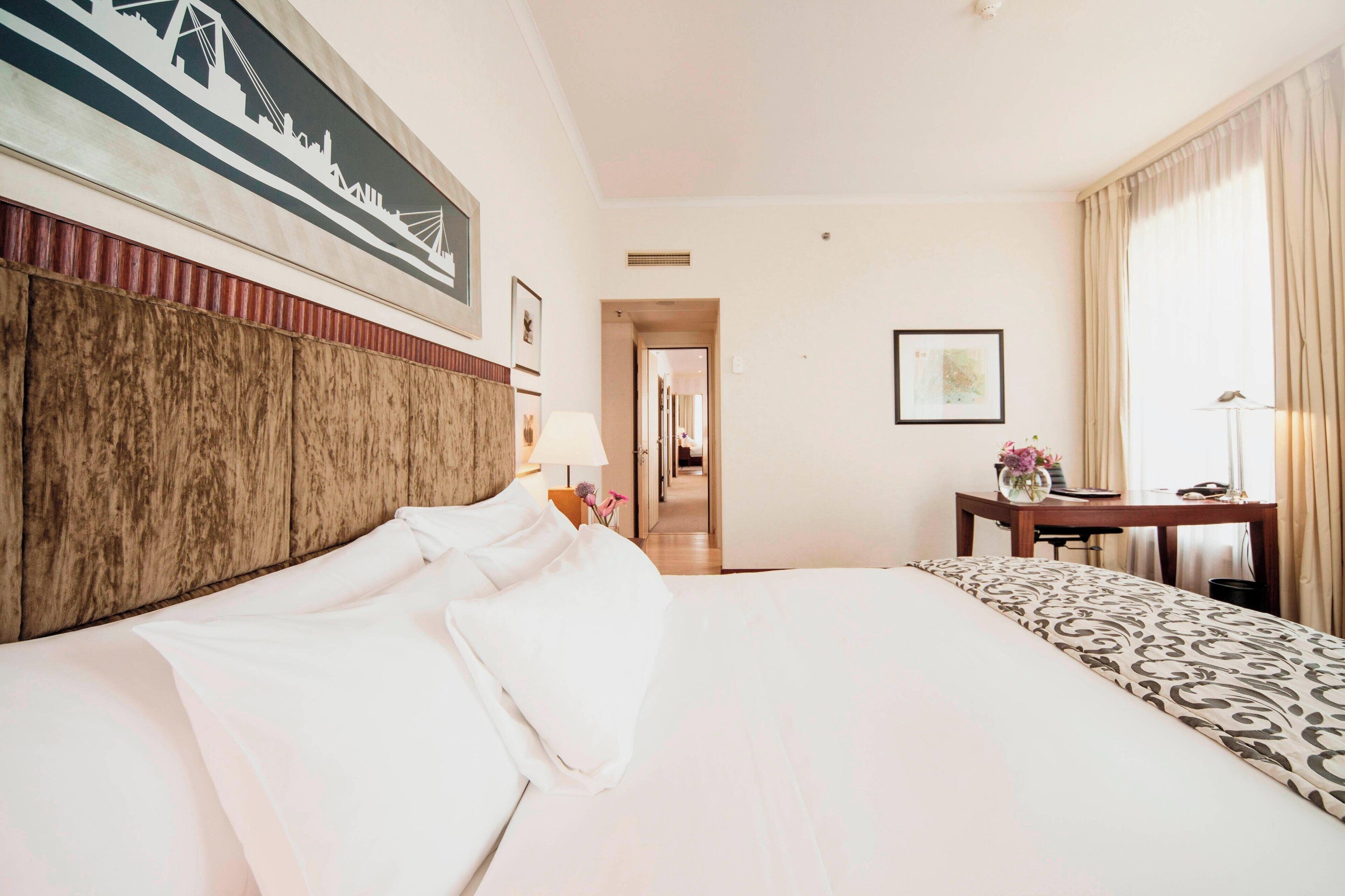 A hotel room with a large bed, a desk, and a chair. There is a framed picture on the wall and a lamp on the desk.