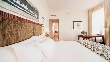 A hotel room with a large bed, a desk, and a chair. There is a framed picture on the wall and a lamp on the desk.