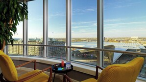 Una habitación con ventanas amplias, dos sillas amarillas, una mesita con una bebida y un libro, y vista a un paisaje urbano.