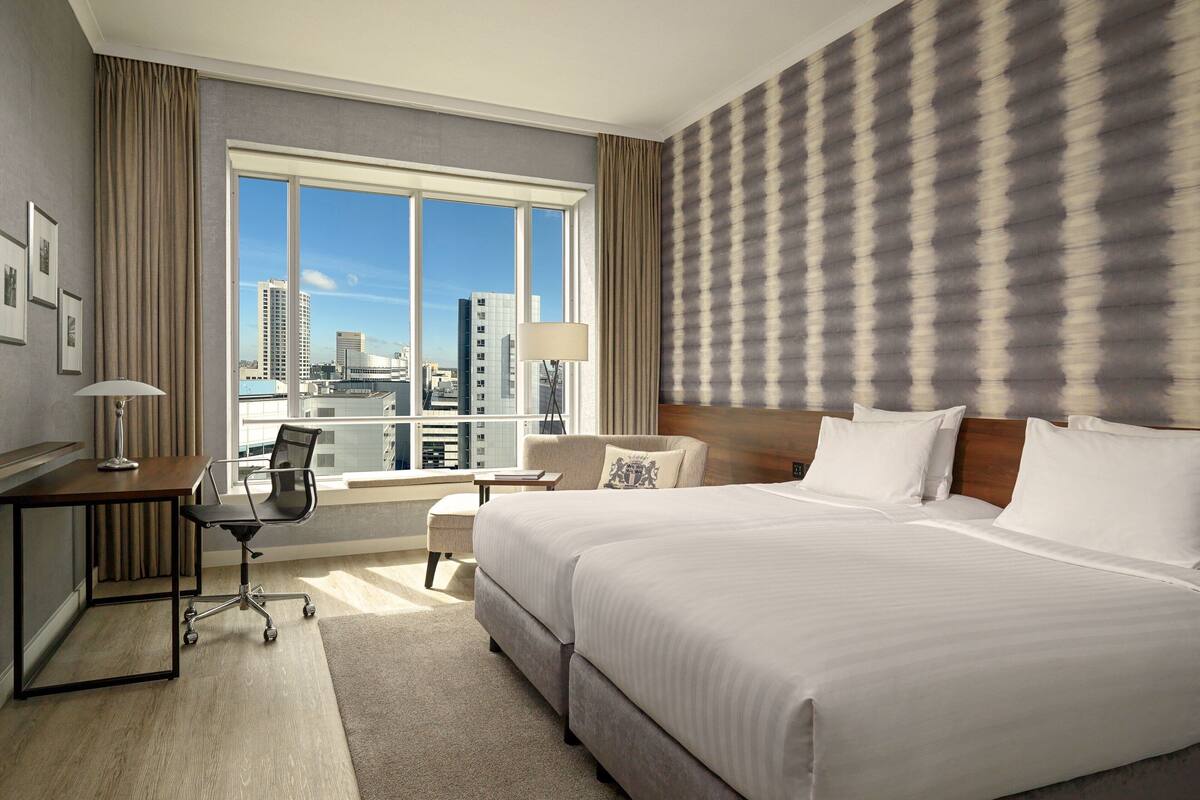 Habitación de hotel con una cama grande, un escritorio, una silla y vistas de la ciudad a través de un ventanal.