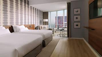 Quarto de hotel com duas camas, uma escrivaninha, uma cadeira, televisão e uma janela grande com vista para a cidade.
