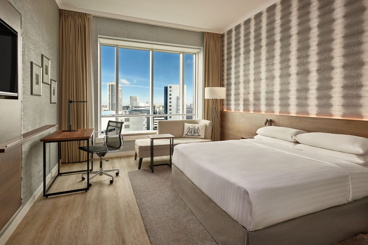 Una habitación de hotel con una cama grande, un escritorio, una silla, un ventanal con vista a la ciudad y una pared con un diseño texturizado.