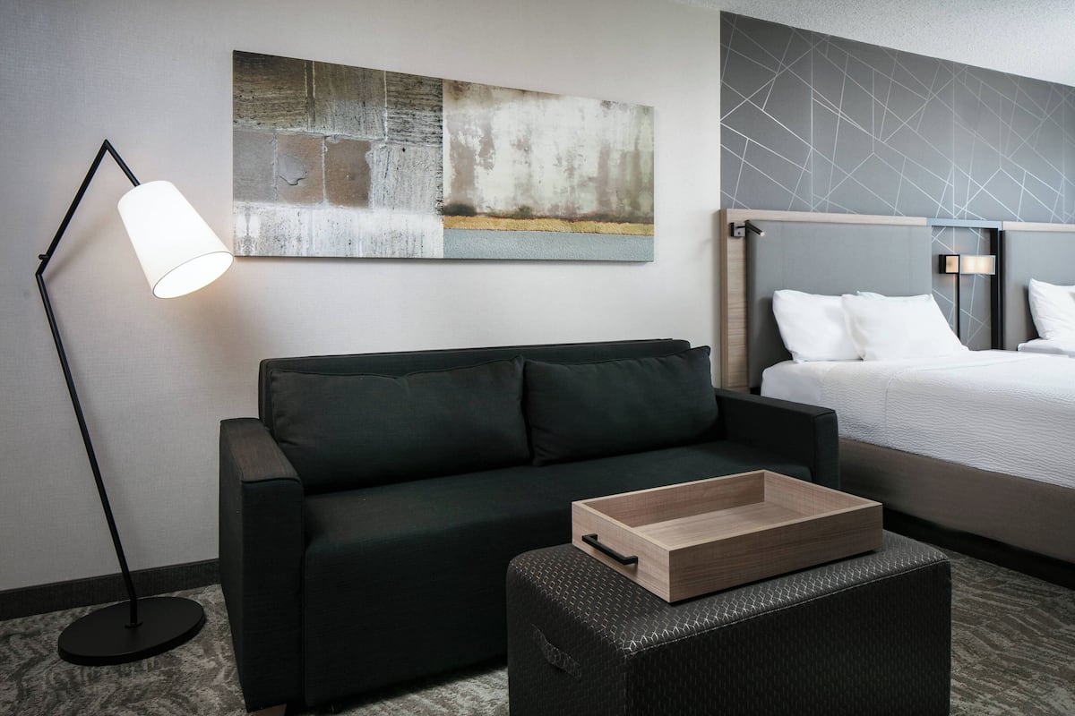 A modern hotel room with a dark-colored sofa, a wooden side table, a bed with white bedding, a floor lamp, and a large abstract painting on the wall.