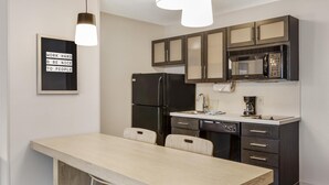 Una cocina moderna con armarios oscuros, un refrigerador negro, un microondas y una cafetera. Hay un comedor de tonos claros con dos sillas.
