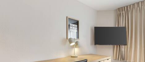 A modern hotel room with a wooden desk, a black chair, a desk lamp, a flat-screen TV, and a mirror.
