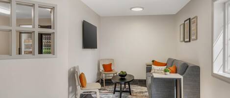 A modern living room with a grey sofa, a small round table, and a white armchair. There is a flat-screen TV mounted on the wall and a window with blinds.