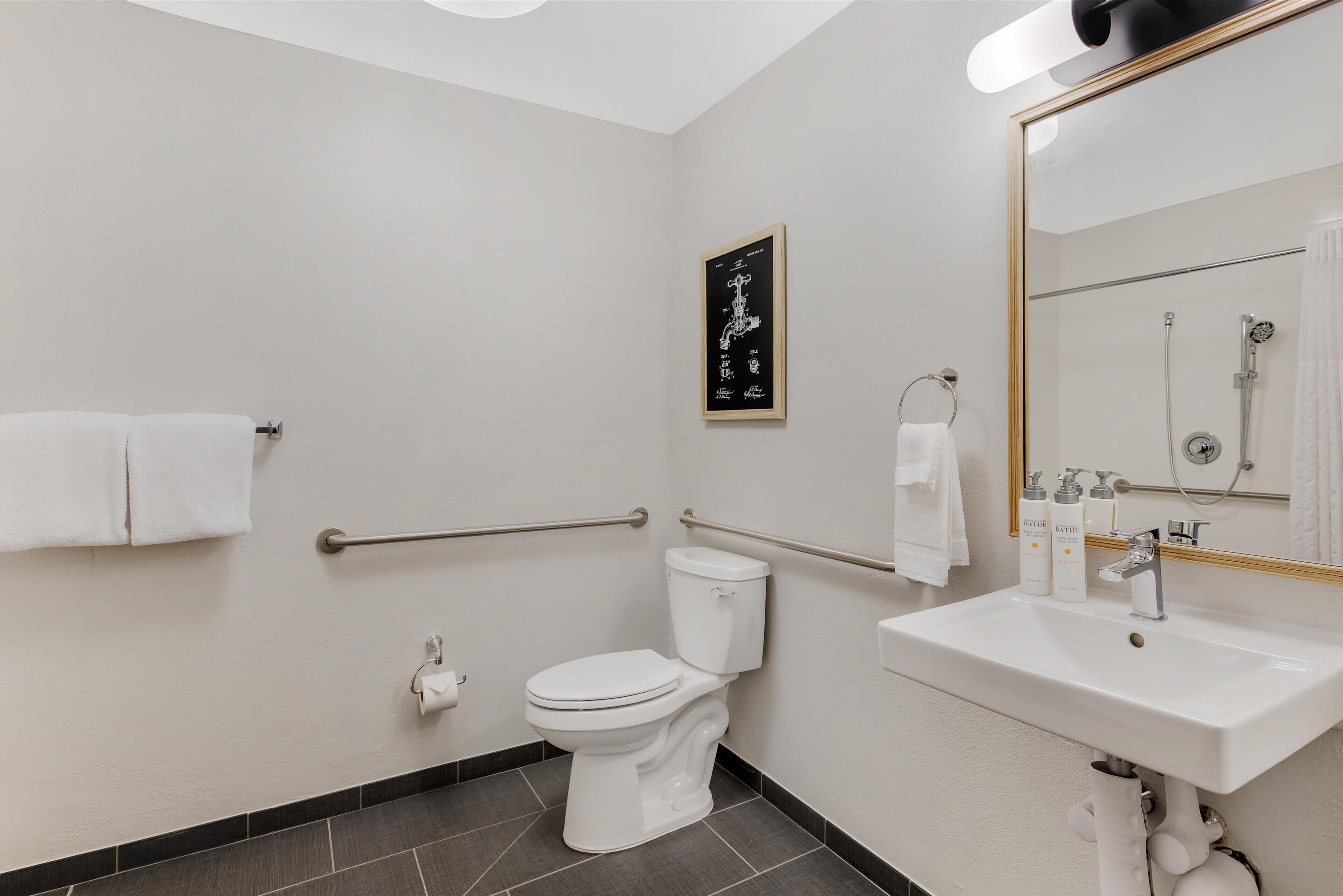 Une salle de bain moderne avec des toilettes blanches, un lavabo équipé de produits de toilette, une douche et des barres d’appui.