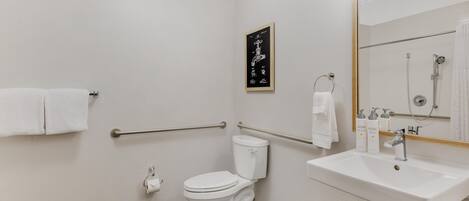 Une salle de bain moderne avec des toilettes blanches, un lavabo équipé de produits de toilette, une douche et des barres d’appui.