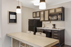 Una cocina moderna con armarios oscuros, un refrigerador negro, un microondas y una cafetera. Hay un comedor de tonos claros con dos sillas.