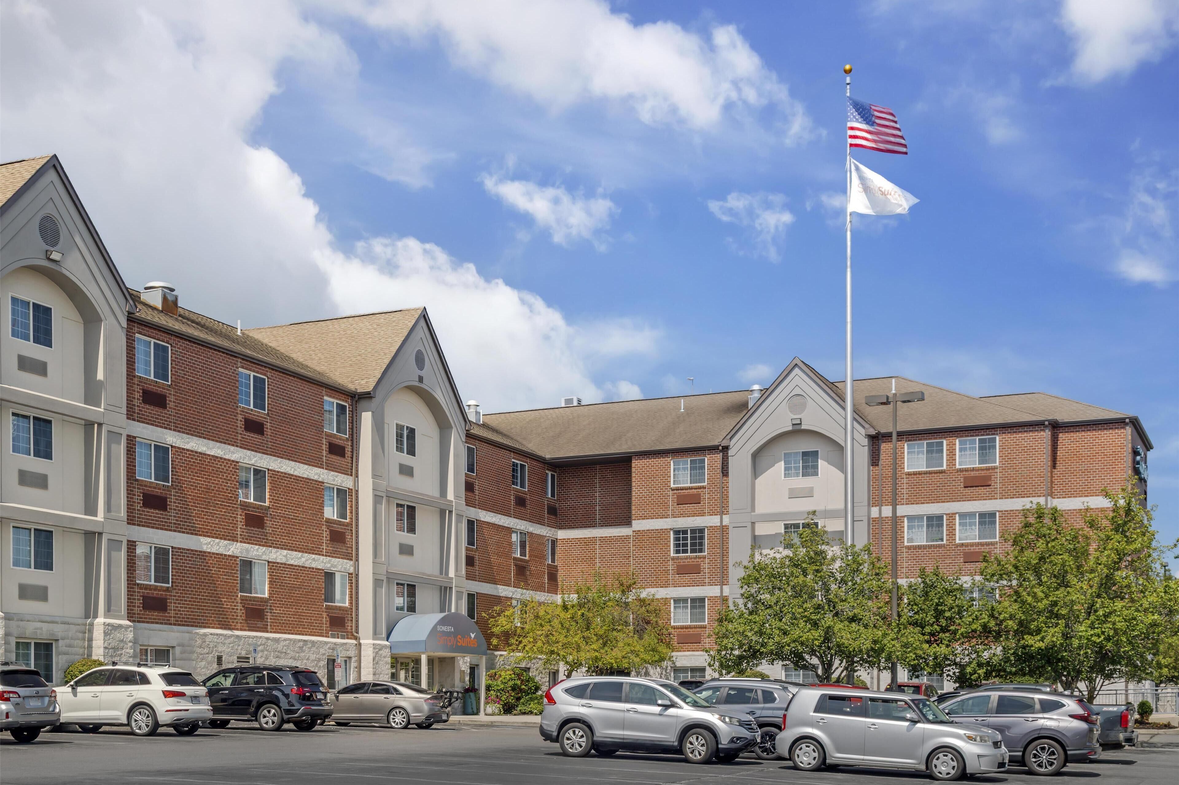 A multi-story hotel with a brick facade, a flagpole with the American flag, and a parking lot with several cars.