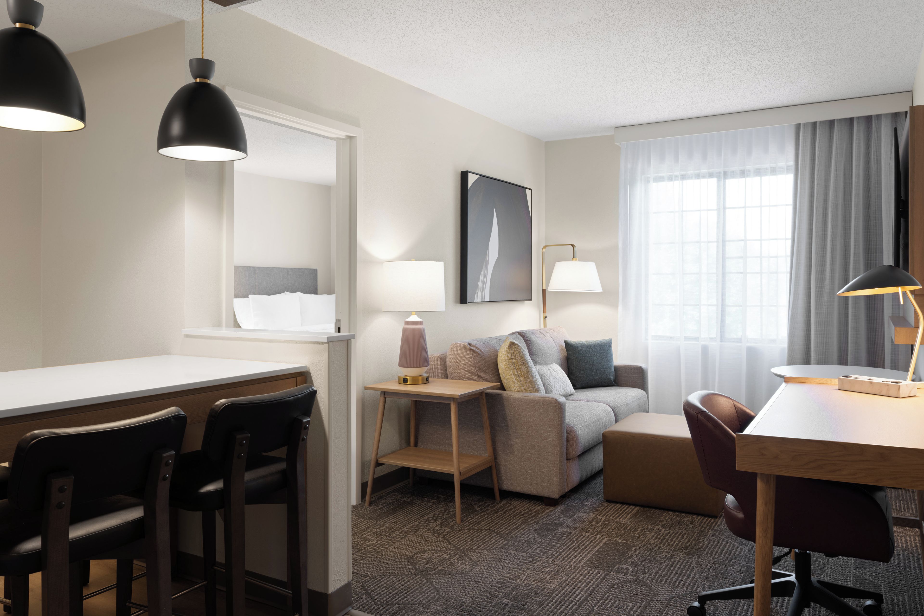 A hotel room with a desk, two chairs, a sofa, a coffee table, a lamp, and a large window with curtains.