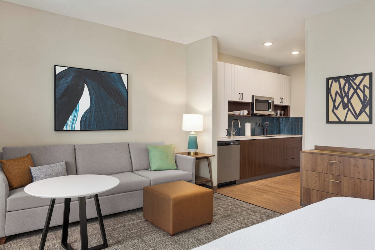 A modern hotel room with a grey sofa, a round white table, a brown ottoman, a bed, a kitchenette with a microwave and stove, and a painting on the wall.