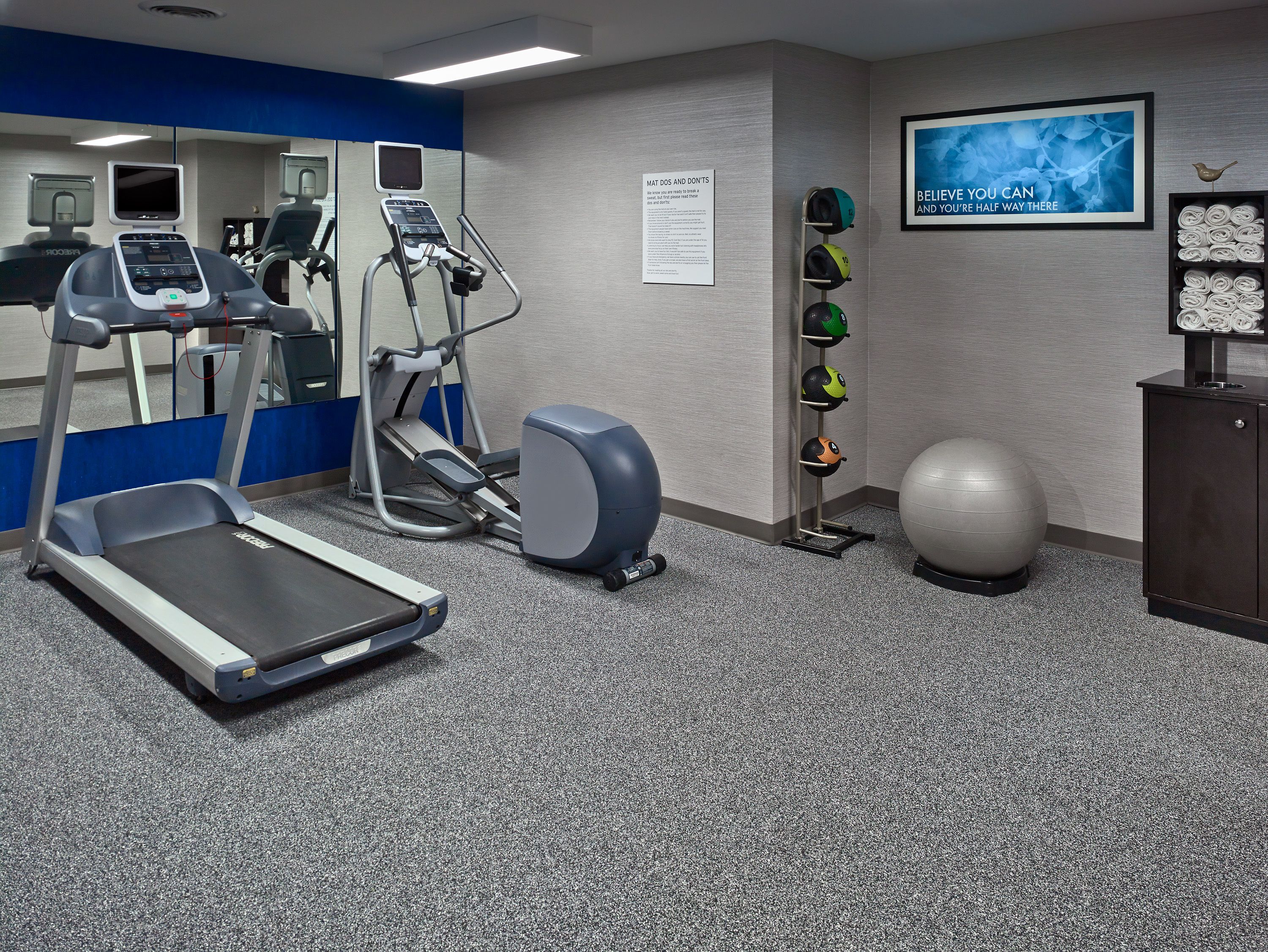 A fitness room with treadmills, an elliptical, a stationary bike, a wall-mounted TV, a motivational poster, a rack of helmets, and a large exercise ball.