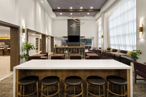 A modern hotel lobby with a central bar area, seating arrangements, and a large television screen. - Staybridge Suites Boston - Burlington by IHG (Burlington)