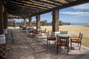 Un patio al aire libre con suelo de piedra, mesas y sillas, con vista a una playa de arena.