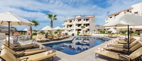 Un área de piscina con sillones de descanso y sombrillas, rodeada de edificios con balcones.