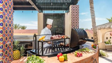 Un chef prépare de la viande sur un barbecue extérieur, entouré de légumes frais, et un menu est écrit sur un tableau noir.