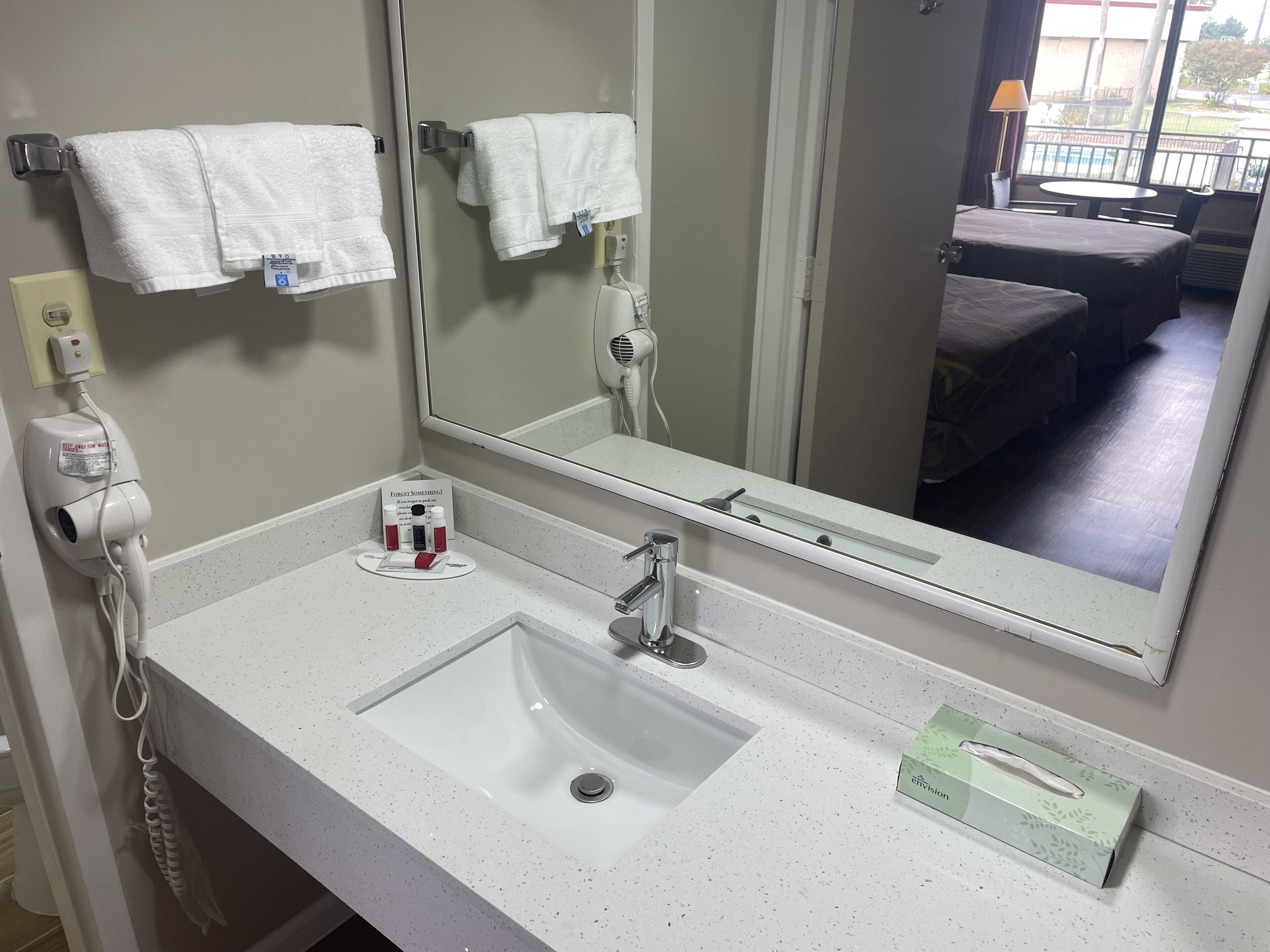 Une salle de bain d’hôtel avec un lavabo blanc, un grand miroir et deux serviettes blanches suspendues à des crochets.